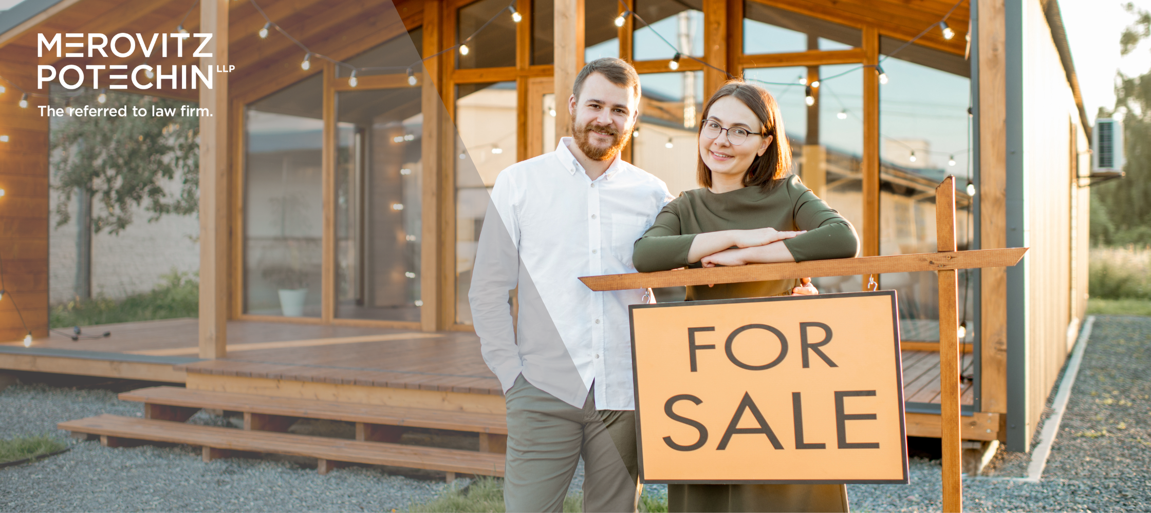 Smiling couple standing in front of a modern wooden house with a 'For Sale' sign, highlighting the importance of spouse consent in real estate transactions, provided by Merovitz Potechin LLP