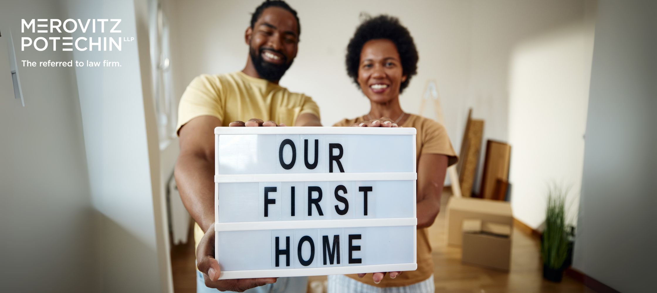 A happy couple holding a sign that says "Our First Home" while standing in their new home. They are smiling, with moving boxes and home decor in the background. The image includes the Merovitz Potechin LLP logo with the tagline "The referred to law firm." This represents the excitement of first-time homebuyers while highlighting the importance of understanding hidden costs homebuyers may face during the purchasing process.